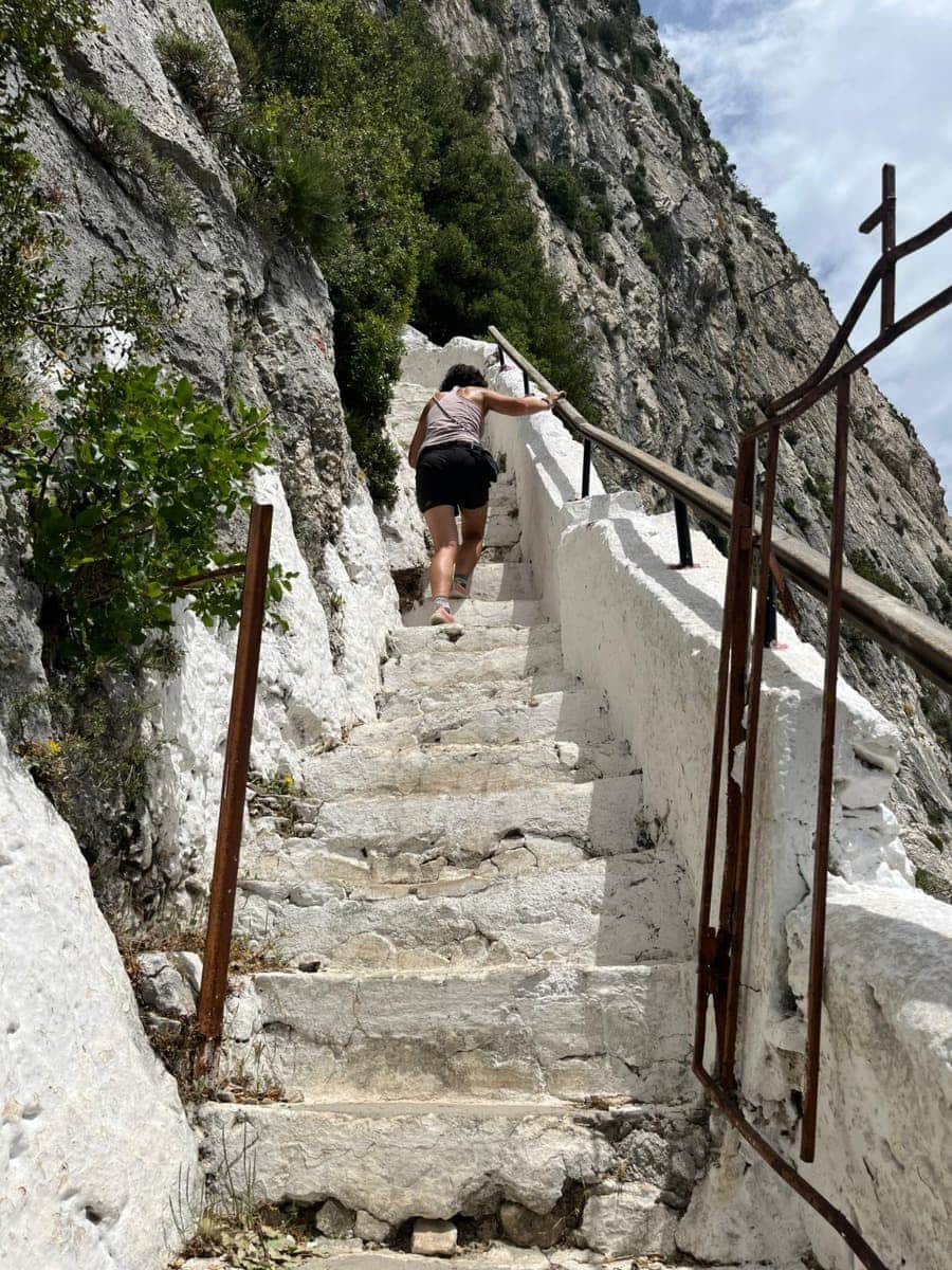 Linda steigt eine steile, in Fels gehauene Steintreppe hinauf, die von einem Geländer gesäumt wird und sich entlang einer Felswand windet. Diese Treppe gehört zum anspruchsvollen Aufstieg zur Höhle des Pythagoras auf Samos.