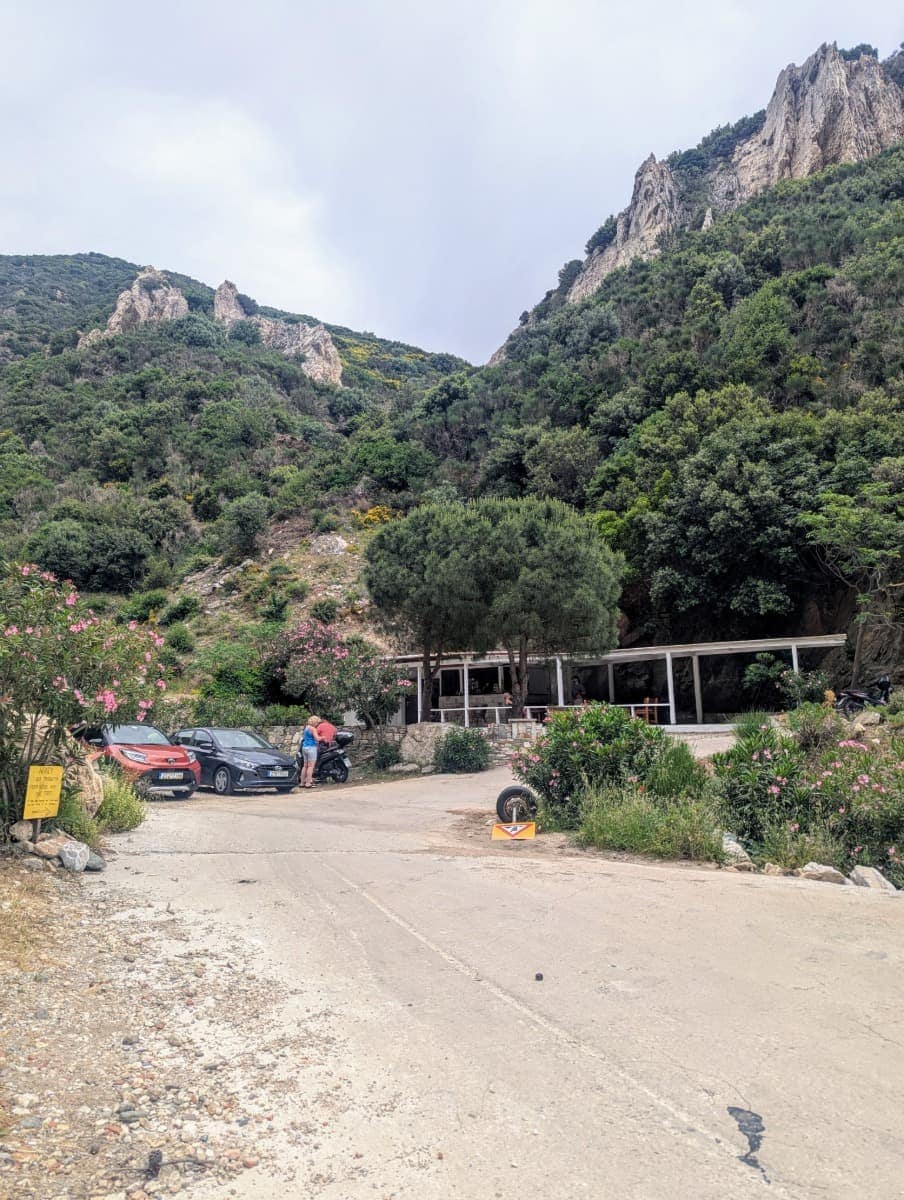 Ein kleiner Parkplatz mit mehreren Autos und einer Taverne liegt eingebettet in bewaldetes Berggelände am Fuß steiler Felsformationen. Von hier aus starten die meisten Besucher ihre Wanderung zur Höhle des Pythagoras.