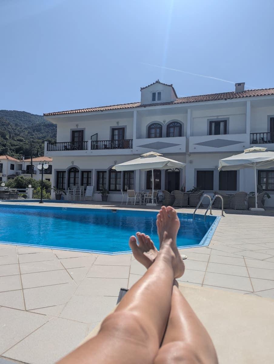 Linda liegt entspannt auf einer Sonnenliege am Pool vor einem weißen zweistöckigen Hotelgebäude mit Balkonen und Sonnenschirmen. Die Iro Residence auf Samos bietet die perfekte Kombination aus Erholung und zentraler Lage für Wanderfreunde.