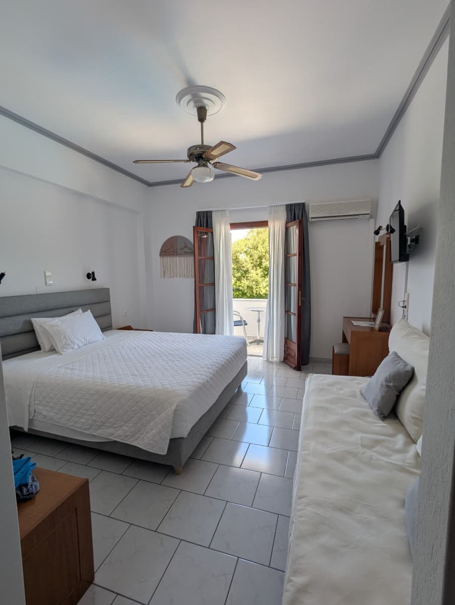 Helles Doppelzimmer im Iro’s Residence Hotel mit Zugang zum Balkon und Blick ins Grüne – ein gemütlicher Rückzugsort beim Urlaub auf Samos. Das Zimmer ist modern eingerichtet mit Deckenventilator, Klimaanlage und Sofa.