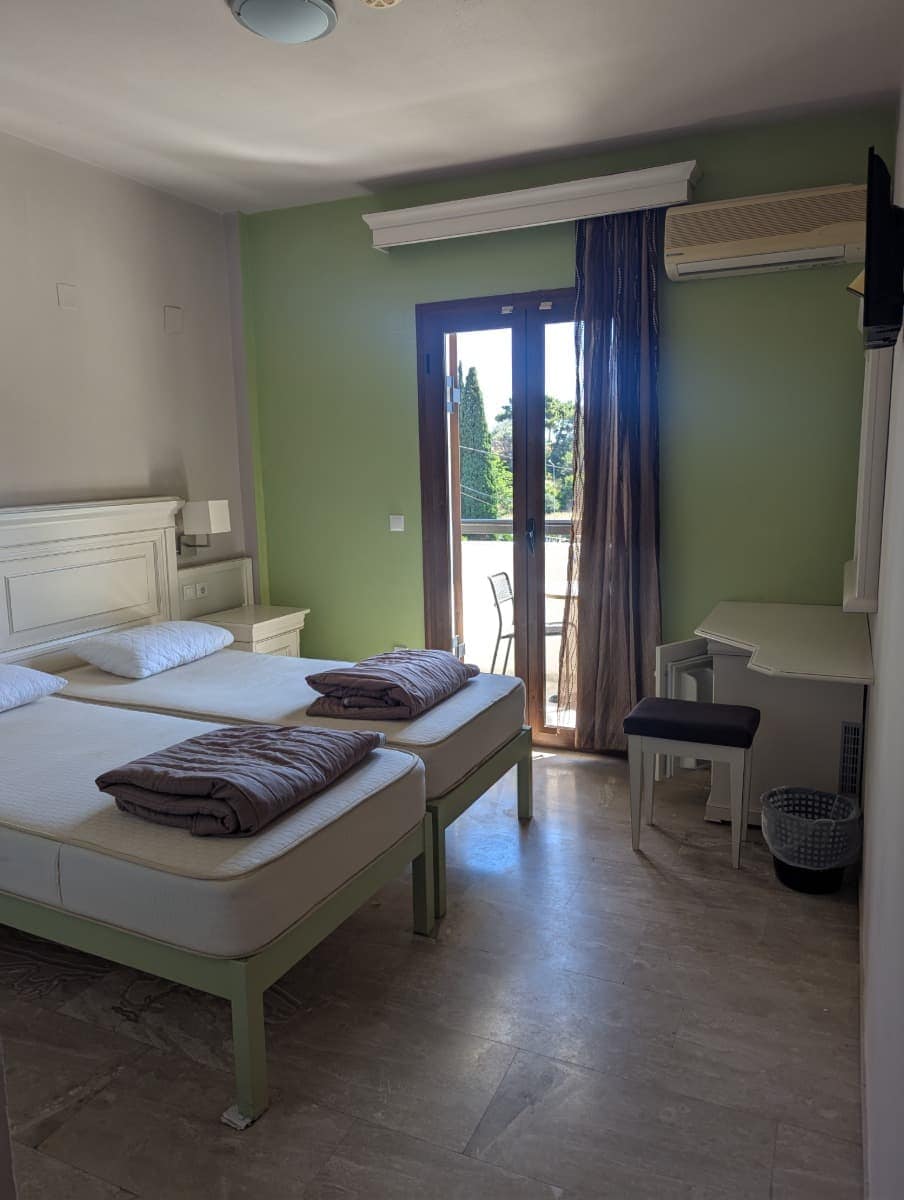 Schlichtes Zweibettzimmer im Sportclub Helen Yolanda mit grün gestrichener Wand, Balkonzugang und Klimaanlage – ideal für aktive Reisende beim Urlaub auf Samos mit Frosch Sportreisen. Ein funktionaler Raum zum Entspannen nach einem Tag voller Inselabenteuer.