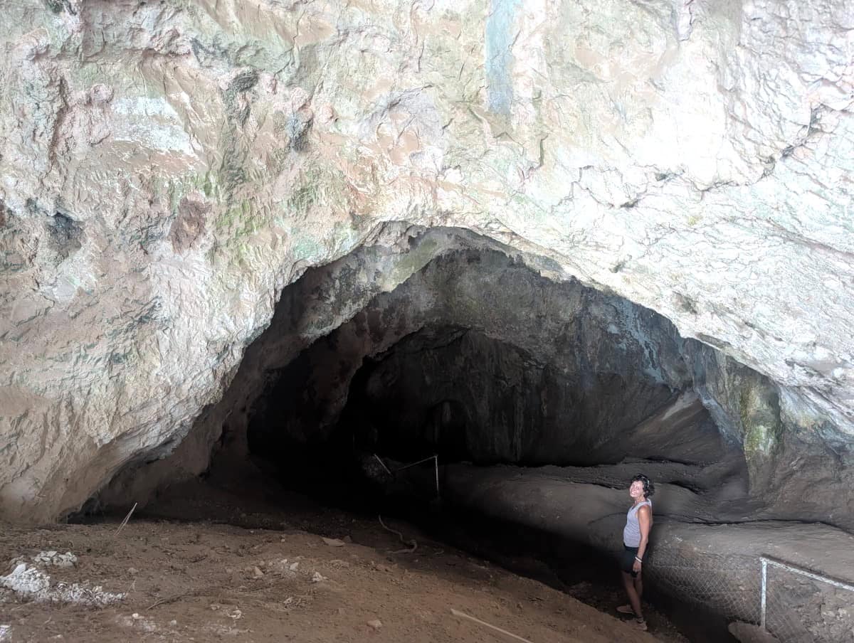 Linda steht vor einem riesigen Höhlengewölbe mit bizarren Felsformationen und einem dunklen Höhleneingang im Hintergrund. Diese benachbarte Höhle befindet sich in unmittelbarer Nähe zur berühmten Höhle des Pythagoras und ist Teil des historischen Geländes.