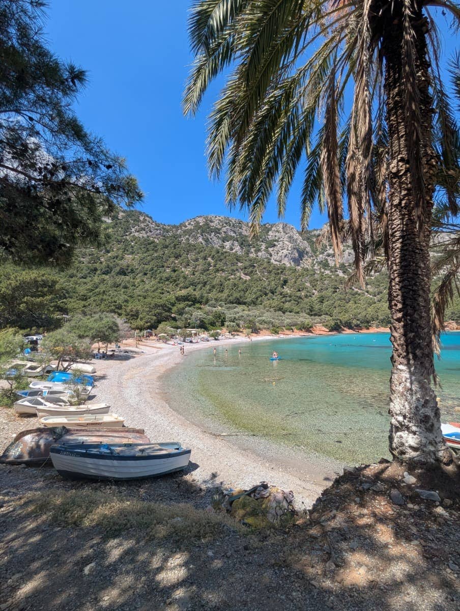 Ein ruhiger Kiesstrand mit türkisfarbenem Wasser liegt eingebettet zwischen grün bewaldeten Hügeln, mit kleinen Booten und vereinzelten Badenden. Der abgelegene Mourtia Beach ist ein echtes Highlight auf Samos für alle, die Natur, Ruhe und kristallklares Wasser suchen.