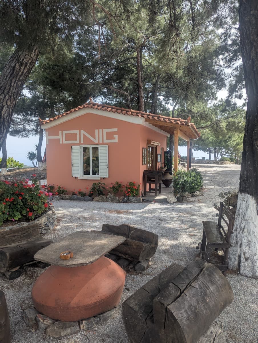 Ein kleines rosafarbenes Häuschen mit der Aufschrift „Honig“ steht idyllisch unter Pinien, umgeben von rustikalen Holzbänken und Blumen. Dieses charmante Verkaufshäuschen ist eine Anlaufstelle für Samos Spezialitäten und bietet lokalen Honig inmitten mediterraner Natur.