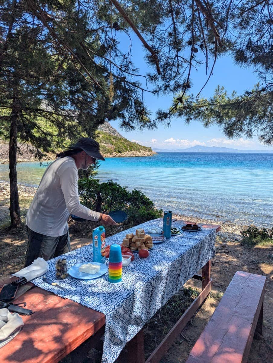 Ein Mann bereitet an einem Holztisch unter Pinien eine Picknickpause mit regionalen Snacks und Meerblick vor, das türkisfarbene Wasser glitzert im Hintergrund. Die abgeschiedene Mikri Lakka Bucht ist ein Geheimtipp auf Samos und perfekt für einen Zwischenstopp bei einer Kajaktour.