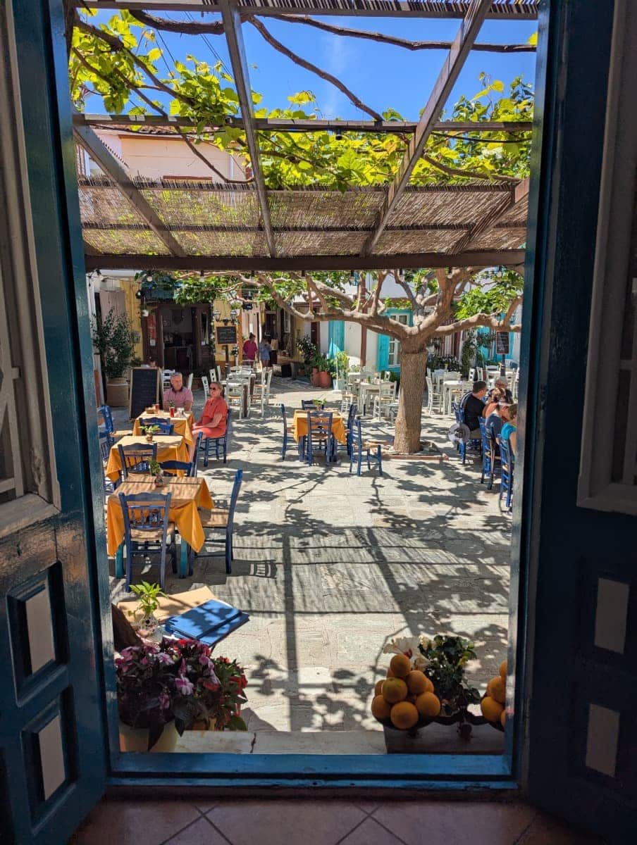 Blick durch eine offene Tür auf einen sonnigen Dorfplatz mit bunten Stühlen und gelb gedeckten Tischen unter einem Weinspalier. Das traditionelle Bergdorf Vouliotes lädt beim Wandern auf Samos zur Rast in urigen Tavernen ein.