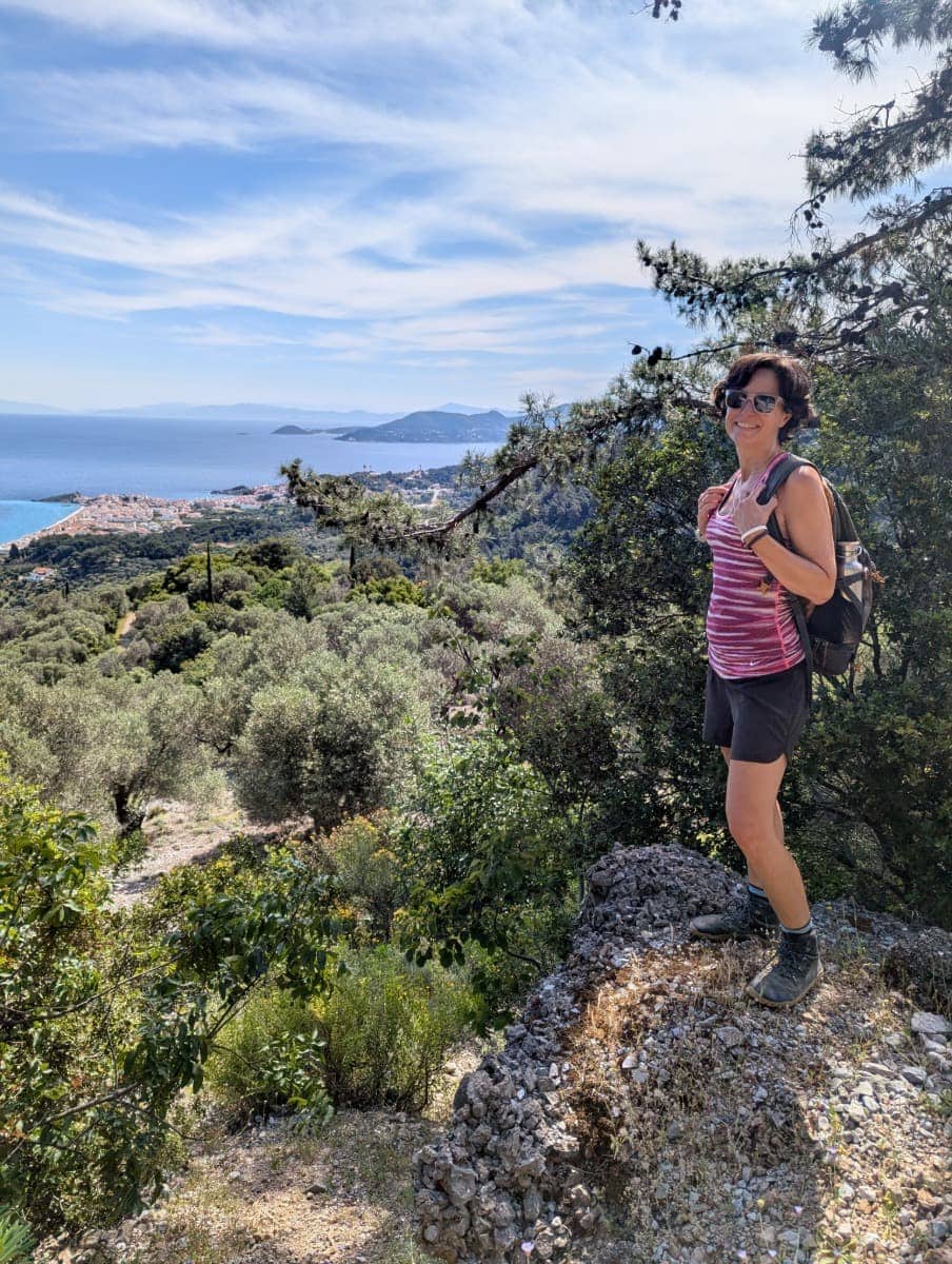 Linda steht auf einem Felsen und genießt den Ausblick auf das Meer und das Dorf Kokkari, eingerahmt von Olivenbäumen und Nadelwald. Der Ort ist ein beliebter Ausgangspunkt zum Wandern auf Samos, besonders auf Panoramawegen.