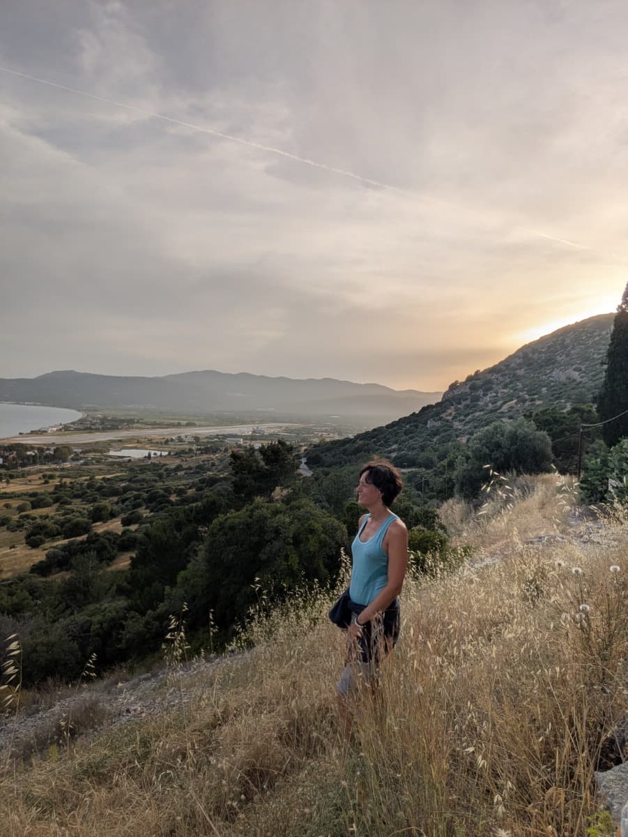 Linda steht auf einer Anhöhe im Abendlicht und blickt über die weite Ebene mit dem Flughafen und der Küste im Hintergrund. Diese Szenerie zeigt die ruhigen und aussichtsreichen Wanderwege auf der Südseite von Samos.