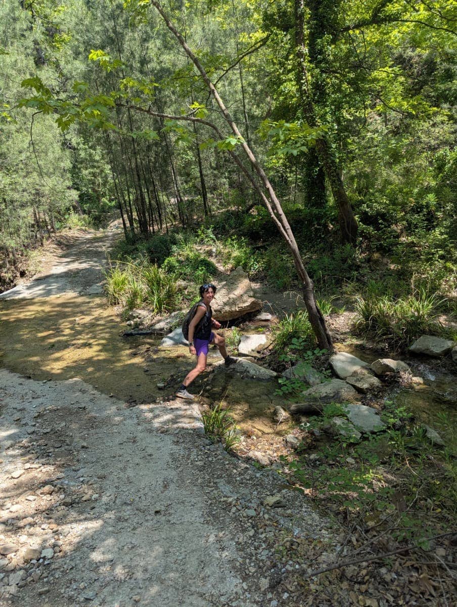Linda überquert einen Bachlauf auf einem schmalen Waldweg im Halbschatten dichter Vegetation. Diese Szene zeigt die idyllischen, naturbelassenen Pfade im Nachtigallental auf Samos.