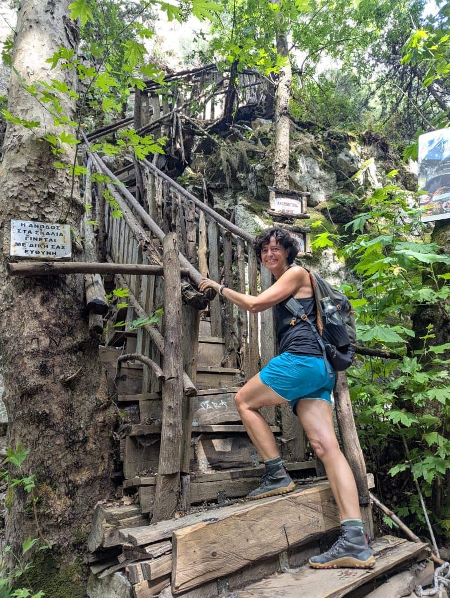 Linda erklimmt eine hölzerne Treppe, die zwischen Bäumen und Felsen zur Taverne Archontissa oberhalb der Potami Schlucht führt. Die Szene zeigt den verwunschenen Charakter dieses Abschnitts oberhalb der Schlucht.