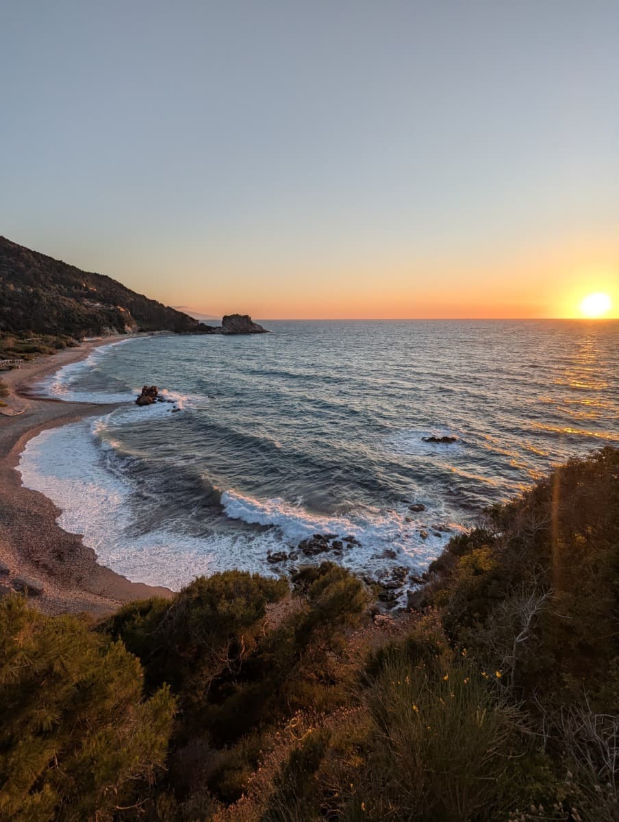 Die untergehende Sonne taucht das Meer und die Felsküste am Potami Beach in warmes Licht, während sanfte Wellen ans Ufer schlagen. Dieser Ort ist ein perfekter Tagesabschluss nach einer ausgedehnten Wanderung auf Samos.