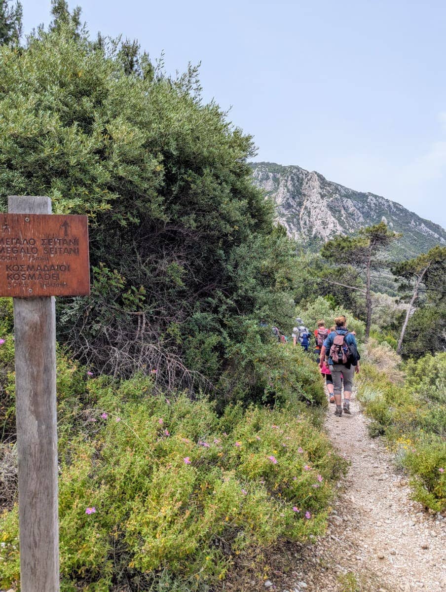 Ein schmaler Wanderpfad führt durch grüne Macchia, begleitet von einer Gruppe Wandernder, neben einem hölzernen Wegweiser mit griechischer und englischer Beschriftung Richtung „Megalo Seitani“. Diese Szenerie zeigt einen typischen Abschnitt beim Wandern auf Samos, wo gut ausgeschilderte Routen durch unberührte Natur zu abgelegenen Stränden führen.