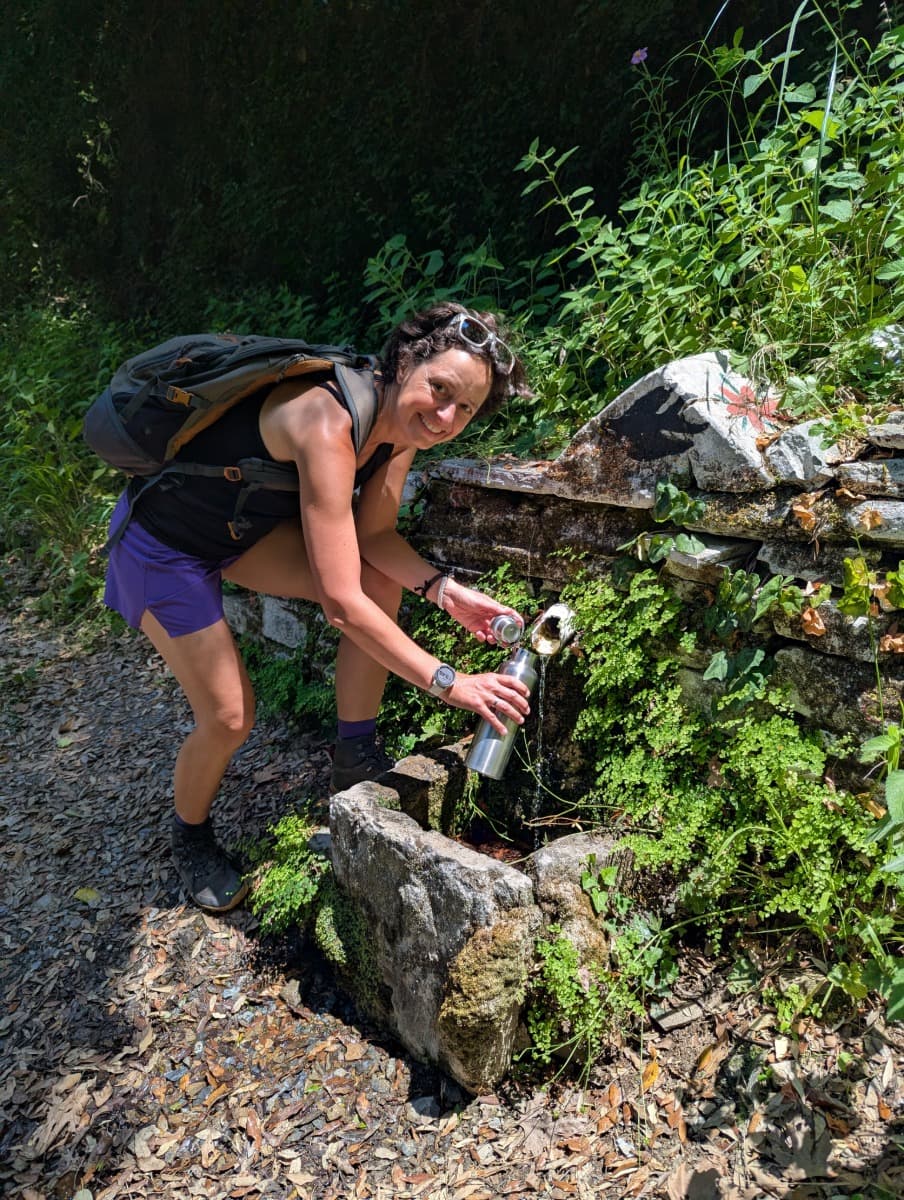 Linda füllt lachend ihre Trinkflasche an einer alten steinernen Wasserquelle, umgeben von Farnen und schattigem Laubwald. Frische Quellen wie diese sind typische Etappen auf den Wanderwegen durch die Wälder von Samos.