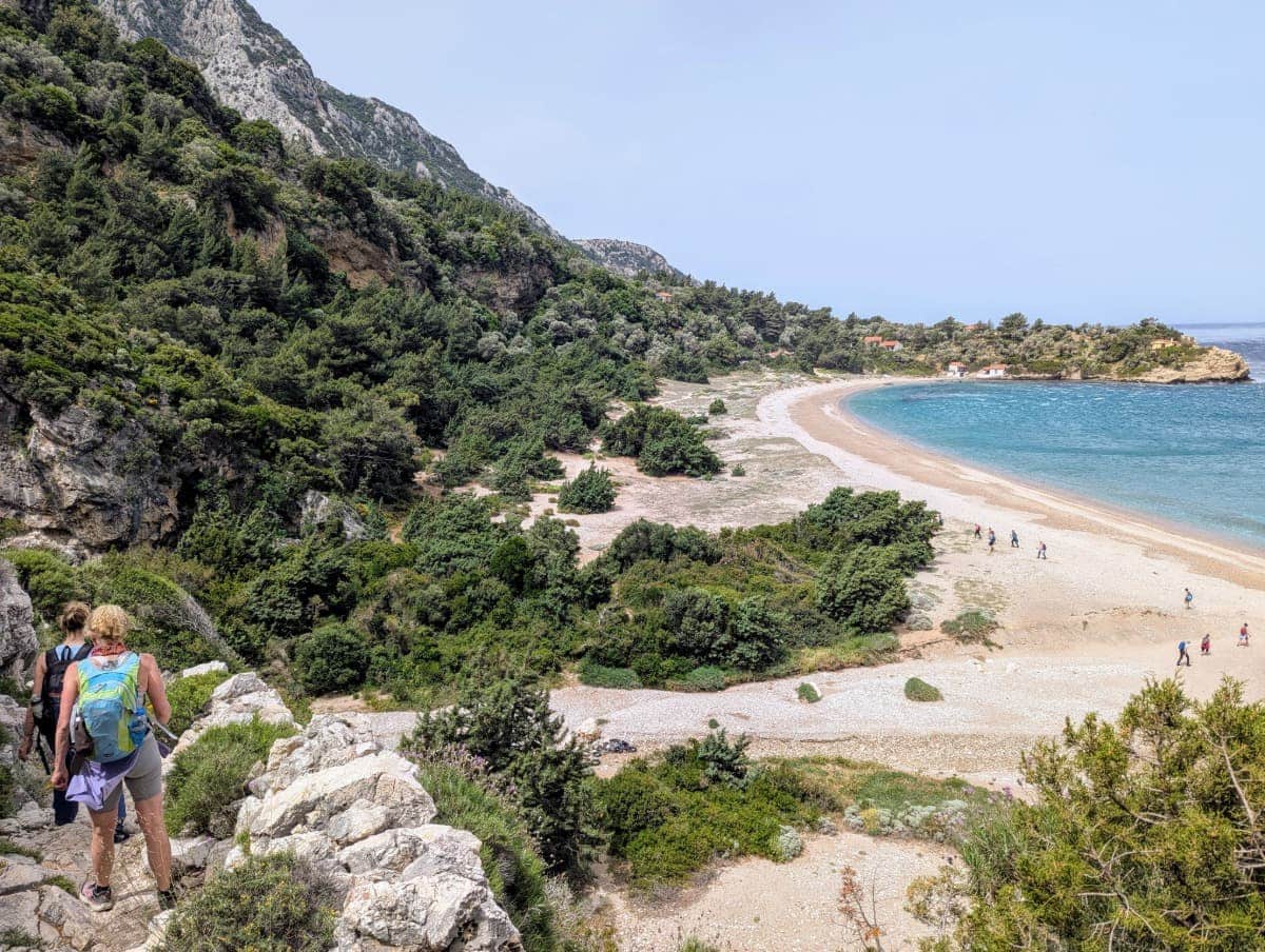 Mehrere Wanderer folgen einem steinigen Pfad oberhalb einer Bucht mit hellen Kiesstränden und türkisblauem Meer. Der Weg zu den Seitani-Stränden gilt als eine der schönsten Küstenwanderungen auf Samos.