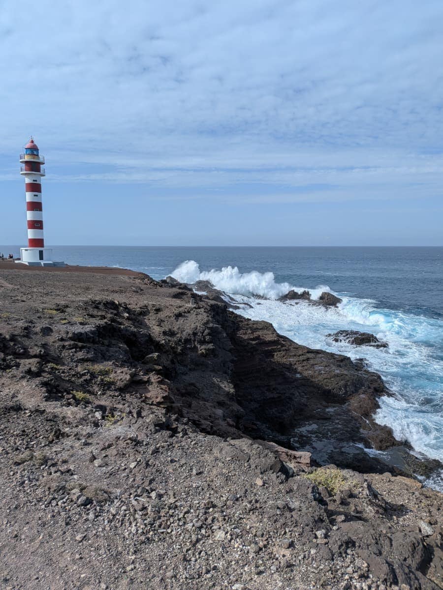 Ein rot-weiß gestreifter Leuchtturm steht auf dunklen Lavaklippen, während Wellen an der Küste von Gran Canaria brechen. Das raue Atlantikpanorama erwartet dich das ganze Jahr über an der Küste.