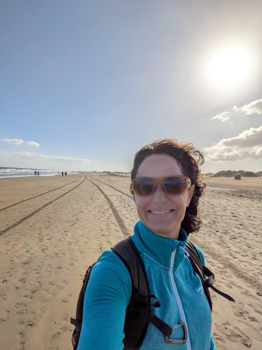 Linda macht ein Selfie an einem breiten Sandstrand mit Fußspuren und wenigen Spaziergängern an der Küste von Gran Canaria. Das windige Wetter verdeutlicht, warum die beste Reisezeit für Gran Canaria ganzjährig sein kann.