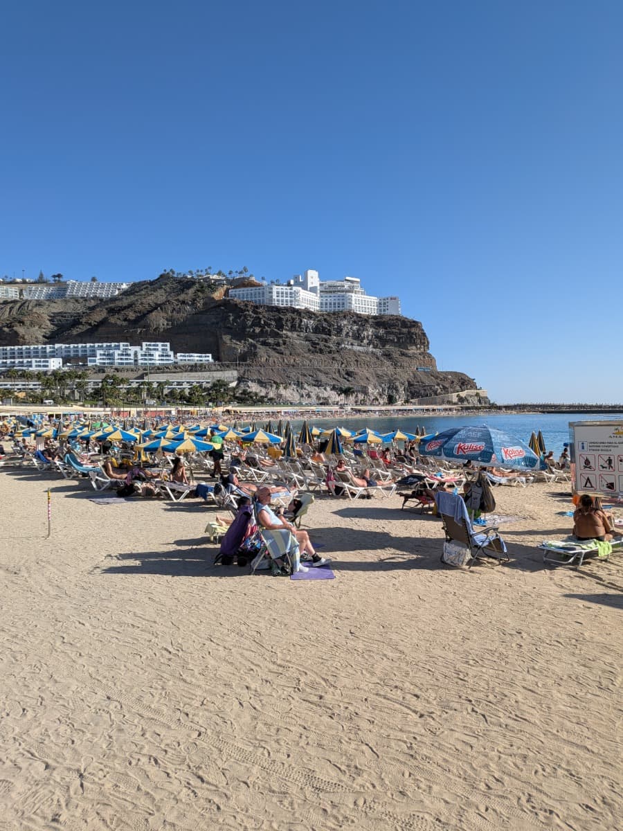 Ein voller Strand mit Liegen und Sonnenschirmen liegt unterhalb einer markanten Felswand und Hotelanlagen auf Gran Canaria. Das Bild zeigt das die besten Reisezeit für Gran Canaria das ganze Jahr über ist wenn man baden möchte.