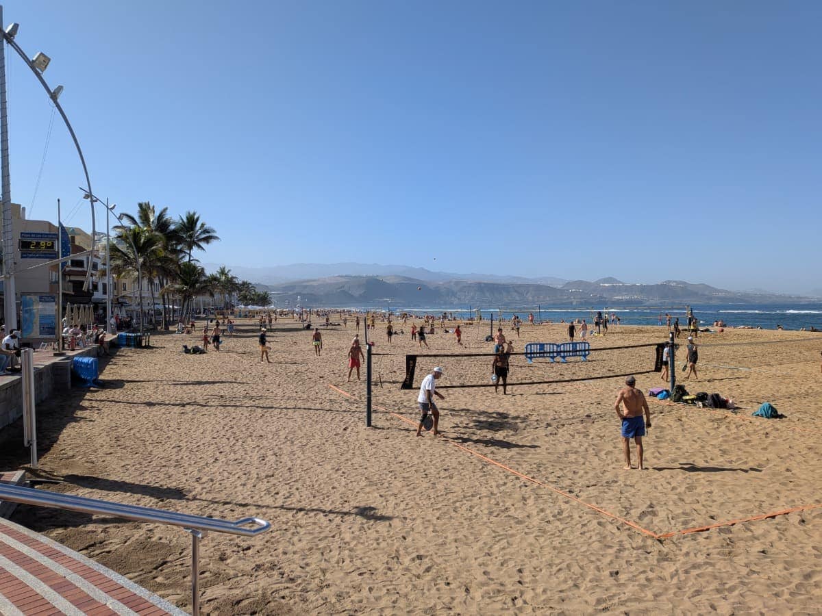 Ein belebter Stadtstrand mit Volleyballfeldern, Palmenpromenade und vielen Menschen an der Küste von Gran Canaria. Die Szene zeigt die warmen Wintermonate auf Gran Canaria.