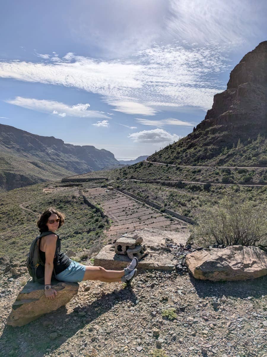 Linda sitzt auf einem Felsen oberhalb eines weiten Tals mit Serpentinenstraße und karger Berglandschaft auf Gran Canaria. Die klare Sicht und das stabile Wetter unterstreichen, warum die beste Reisezeit auf Gran Canaria jederzeit sein kann.