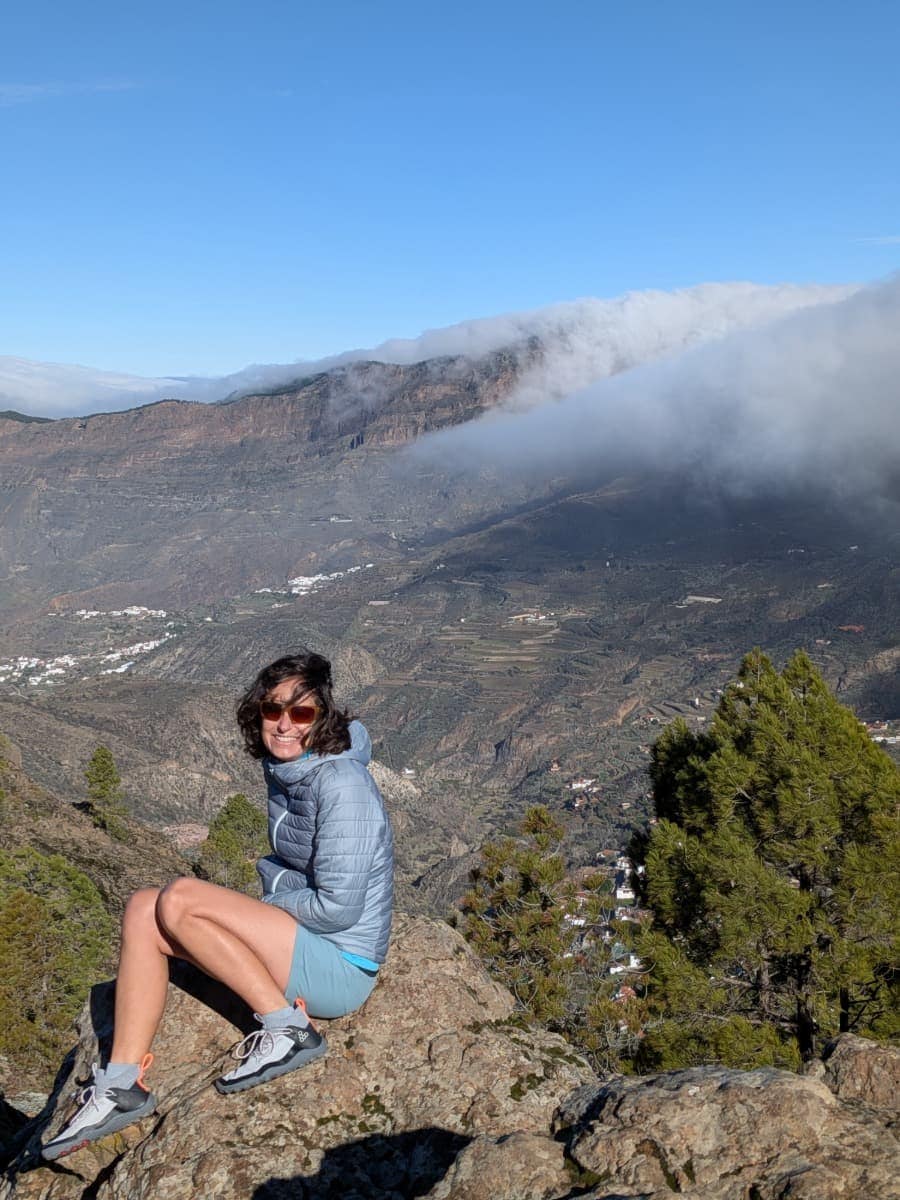 Linda sitzt auf einem Aussichtspunkt mit Blick über ein tiefes Tal, teilweise von Wolken umhüllt, im Hochland von Gran Canaria. Die Mischung aus Sonne und Wolken zeigt die abwechslungsreichen Wetterstimmungen im Winter auf Gran Canaria.