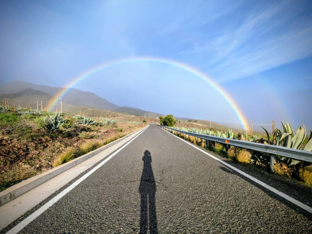 Eine gerade Straße führt durch eine trockene Landschaft, darüber spannt sich ein vollständiger Regenbogen auf Gran Canaria. Das seltene Naturschauspiel zeigt, wie abwechslungsreich auch die beste Reisezeit für Gran Canaria sein kann.